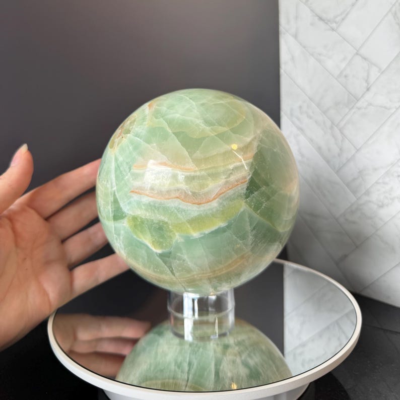 May include: A polished, spherical green onyx stone featuring natural banding in shades of light green, white, and soft orange. The stone is mounted on a clear, circular acrylic stand that rests on a reflective, circular base.