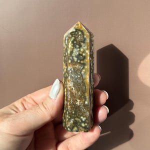 Puede incluir: Una piedra preciosa pulida y columnar con un patr&oacute;n moteado de verde, marr&oacute;n y blanco. El cristal tiene una parte superior puntiaguda y se sostiene en una mano. El fondo es marr&oacute;n liso y apagado.
