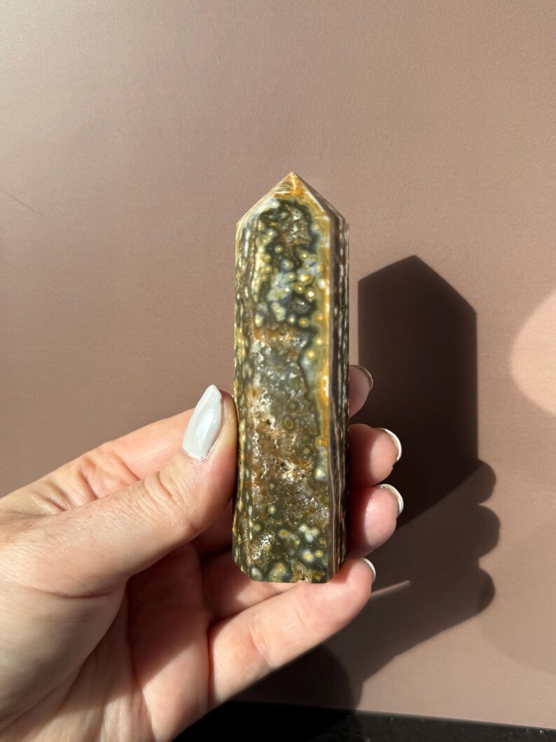 Puede incluir: Una piedra preciosa pulida y columnar con un patr&oacute;n moteado de verde, marr&oacute;n y blanco. El cristal tiene una parte superior puntiaguda y se sostiene en una mano. El fondo es marr&oacute;n liso y apagado.