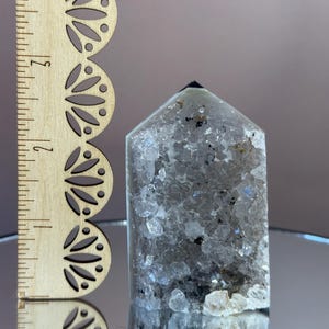 May include: A clear quartz crystal point with internal inclusions and a dark tip. A wooden ruler with decorative cutouts is next to the crystal. The crystal is approximately 3 inches tall.