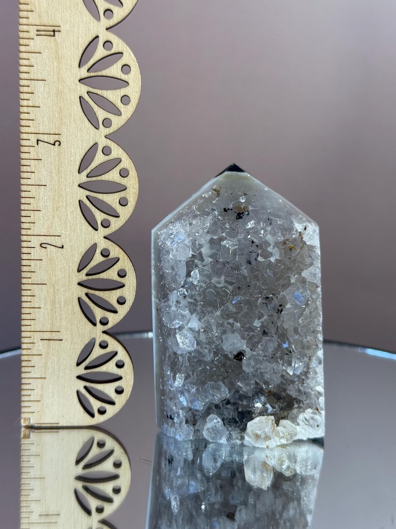 May include: A clear quartz crystal point with internal inclusions and a dark tip. A wooden ruler with decorative cutouts is next to the crystal. The crystal is approximately 3 inches tall.