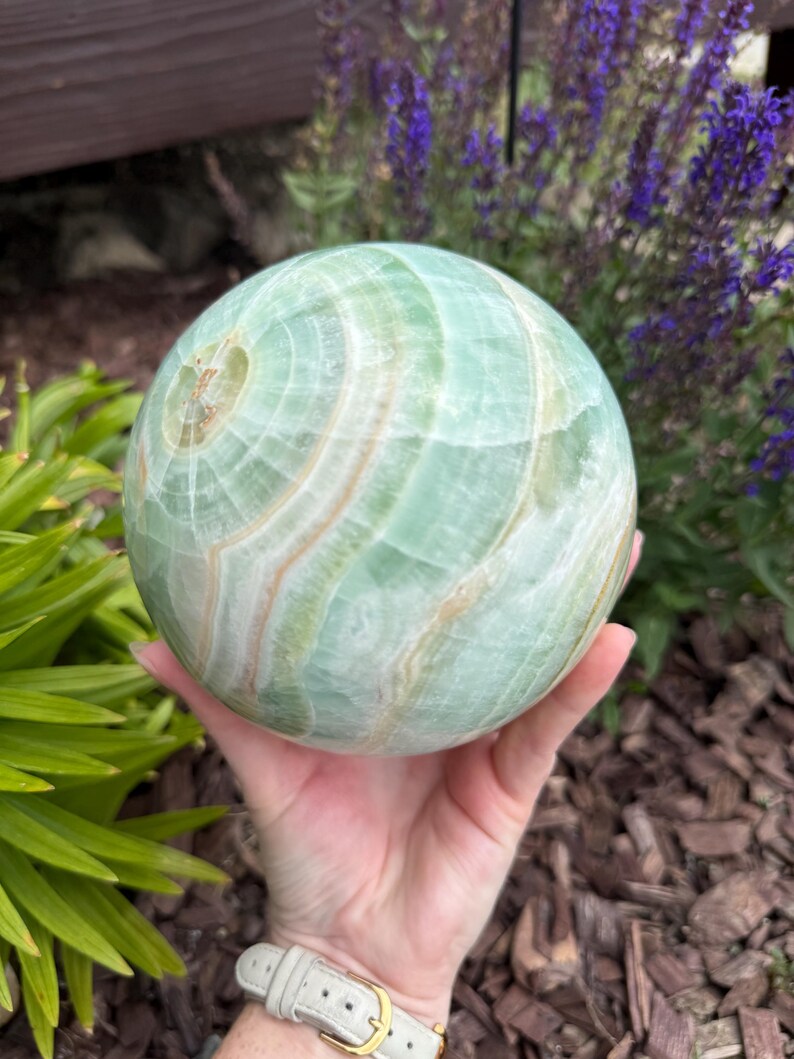 May include: A polished spherical stone featuring a smooth surface with swirling patterns of light mint green, pale cream, and soft white bands. The orb displays natural mineral inclusions and translucent layers throughout its rounded form.
