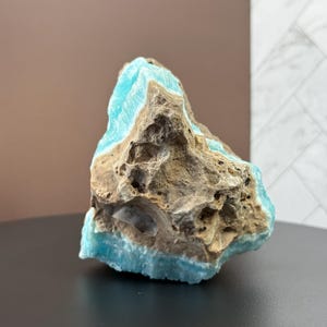 May include: A rough-hewn, natural stone specimen featuring a striking contrast of colors. The stone displays a vibrant turquoise hue along its edges, juxtaposed against a textured, earthy brown and gray core. The mineral sits on a black surface.