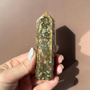 Puede incluir: Un obelisco de piedra preciosa rectangular y pulido. La piedra presenta un patr&oacute;n moteado de colores verde oscuro, marr&oacute;n y crema. La parte superior tiene forma de pir&aacute;mide puntiaguda. El obelisco se sostiene en una mano.