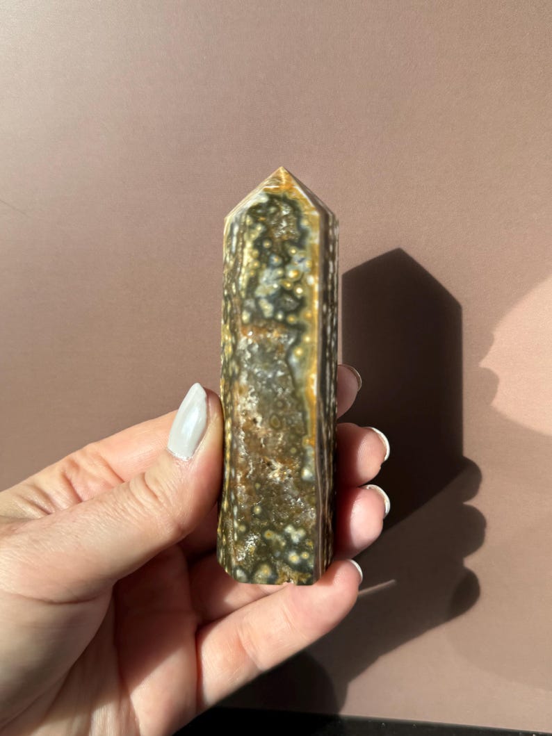 Puede incluir: Un obelisco de piedra preciosa rectangular y pulido. La piedra presenta un patr&oacute;n moteado de colores verde oscuro, marr&oacute;n y crema. La parte superior tiene forma de pir&aacute;mide puntiaguda. El obelisco se sostiene en una mano.