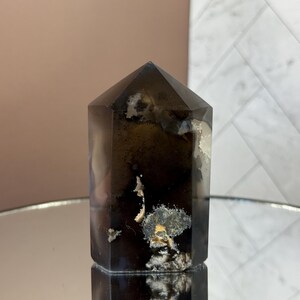 May include: A dark brown crystal tower with a faceted top. The crystal has white and yellow inclusions and sits on a reflective surface. The background is a mix of pink and white.