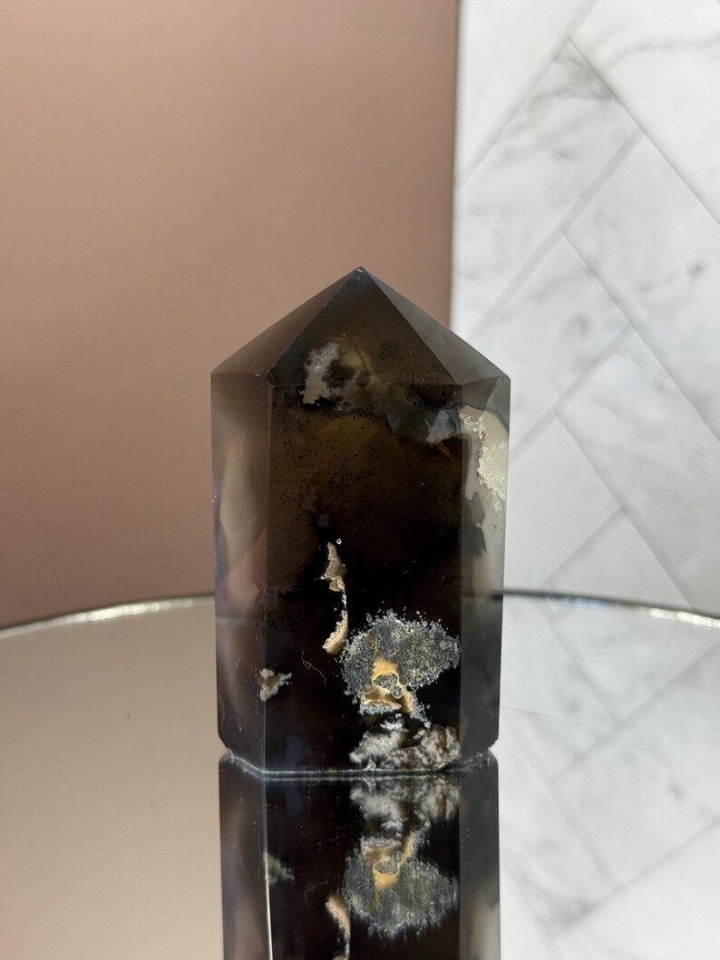 May include: A dark brown crystal tower with a faceted top. The crystal has white and yellow inclusions and sits on a reflective surface. The background is a mix of pink and white.