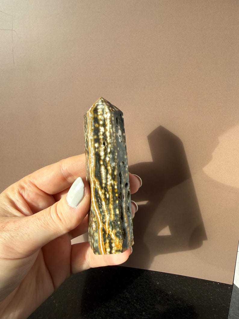 Puede incluir: Un obelisco de piedra preciosa pulida con una parte superior puntiaguda. La piedra presenta una mezcla de colores gris oscuro, marr&oacute;n y crema con un patr&oacute;n moteado. El obelisco se sostiene en una mano sobre un fondo neutro.