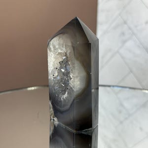 May include: A tall, polished crystal point with a dark gray and white banded pattern. The crystal has a pointed top and a hollowed-out section with sparkling, crystalline formations. The crystal is displayed on a reflective surface.
