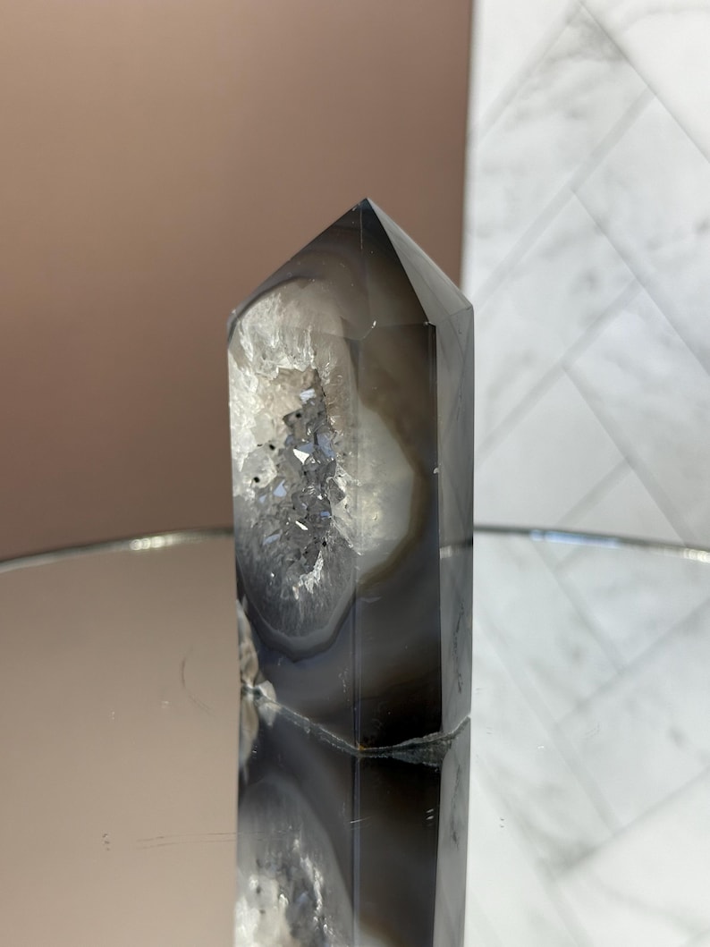 May include: A tall, polished crystal point with a dark gray and white banded pattern. The crystal has a pointed top and a hollowed-out section with sparkling, crystalline formations. The crystal is displayed on a reflective surface.