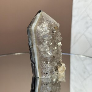 May include: A polished crystal geode with a pointed top. The crystal features bands of gray, white, and black, with a cluster of smaller crystals on one side. The geode is displayed on a reflective surface.
