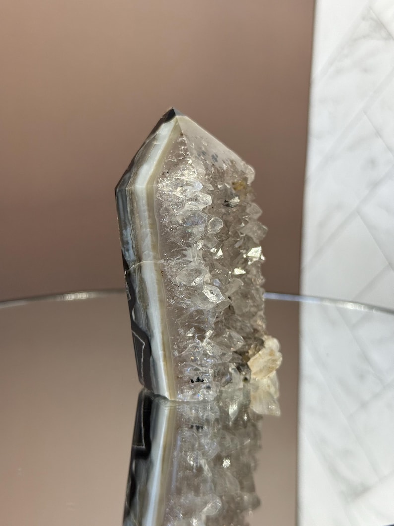 May include: A polished crystal geode with a pointed top. The crystal features bands of gray, white, and black, with a cluster of smaller crystals on one side. The geode is displayed on a reflective surface.