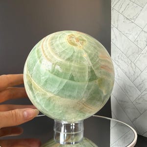 May include: A polished green onyx sphere featuring natural banded patterns in shades of light green, white, and soft orange. The stone orb is displayed on a clear cylindrical stand, showcasing its smooth, reflective surface and intricate mineral layers.