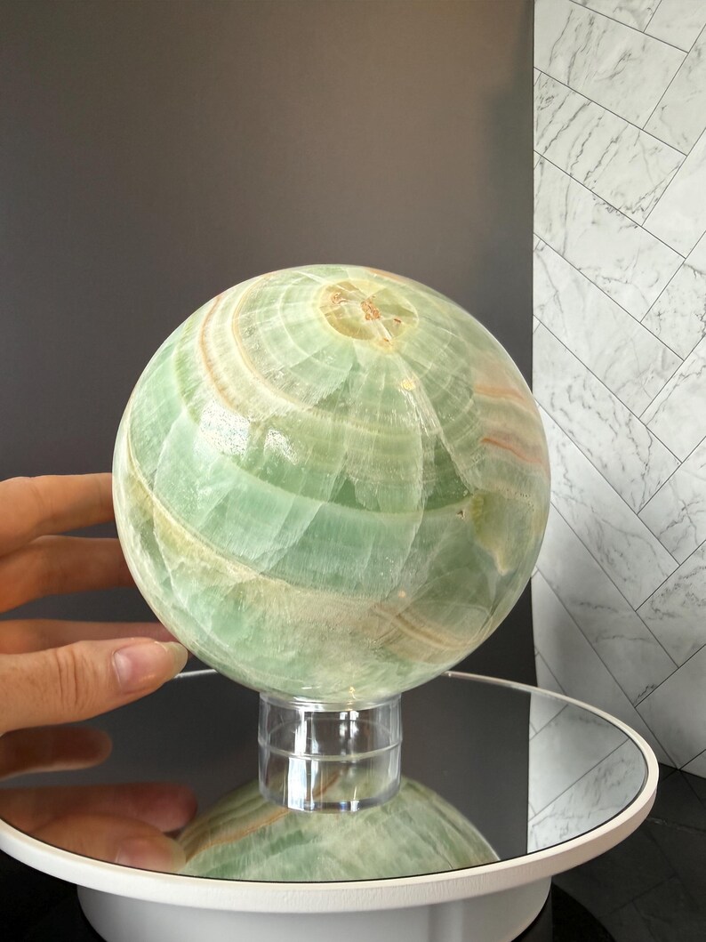 May include: A polished green onyx sphere featuring natural banded patterns in shades of light green, white, and soft orange. The stone orb is displayed on a clear cylindrical stand, showcasing its smooth, reflective surface and intricate mineral layers.