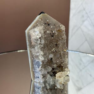 May include: A clear quartz crystal point with a dark tip and a rough, textured surface. The crystal has a light gray and white color with small dark inclusions. The base of the crystal is covered in small, sparkling quartz crystals.