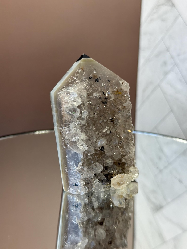 May include: A clear quartz crystal point with a dark tip and a rough, textured surface. The crystal has a light gray and white color with small dark inclusions. The base of the crystal is covered in small, sparkling quartz crystals.