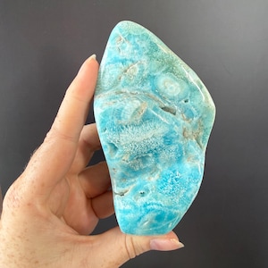 Puede incluir: Una losa pulida de piedra larimar azul con intrincados patrones de remolinos en blanco y azul claro, texturas cristalinas e inclusiones minerales naturales. La piedra tiene una forma orgánica irregular con un acabado superficial liso.