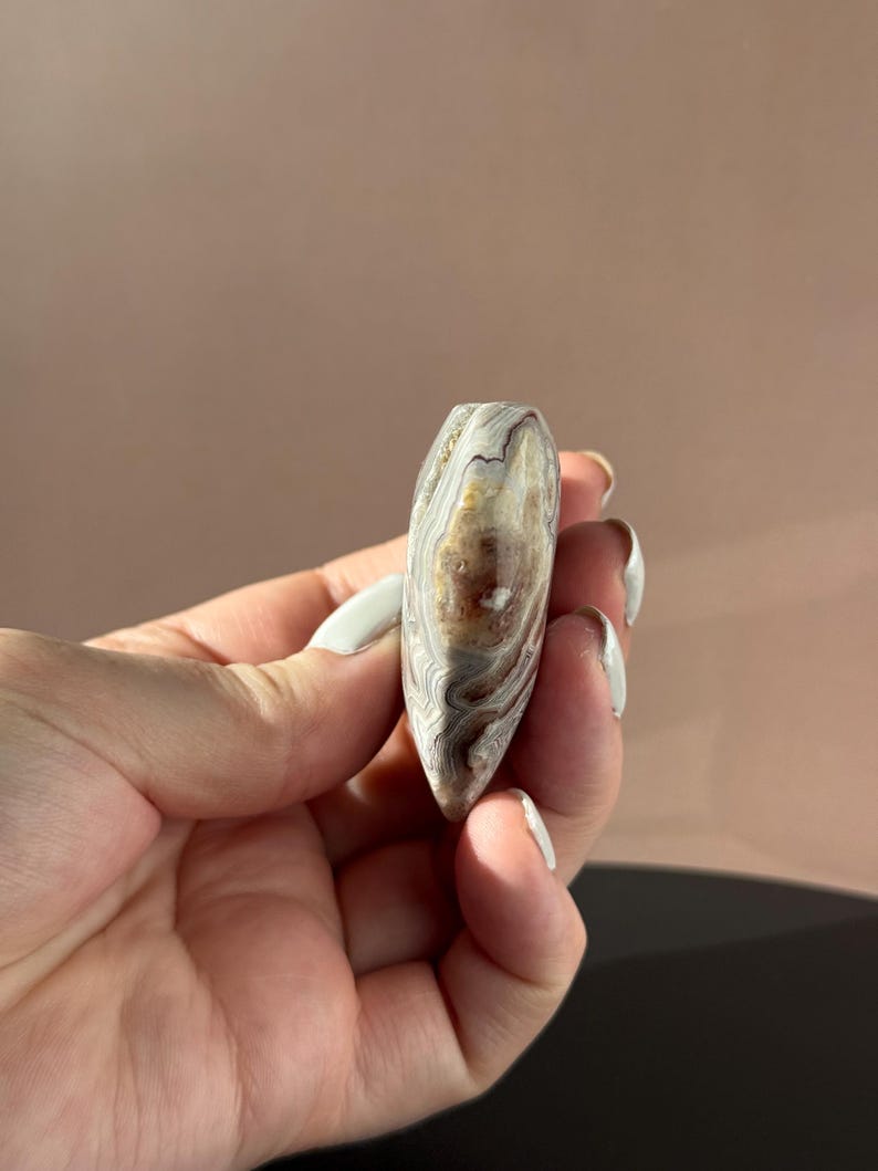 May include: A polished, teardrop-shaped stone with a swirling pattern of brown, cream, and gray bands. The stone is held in a hand with white nail polish. The stone's surface is smooth and reflective, showcasing natural variations in color and texture.