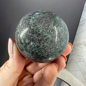May include: A polished, spherical gemstone with a mottled appearance. The stone features a mix of deep green and dark red hues, creating a speckled pattern across its surface. The gemstone is held in a hand, showcasing its smooth, rounded form.