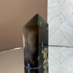 May include: A dark brown crystal obelisk with a pointed top. The crystal has a polished surface with areas of lighter color and some gold-colored inclusions. The obelisk is standing upright, reflecting on a mirrored surface.