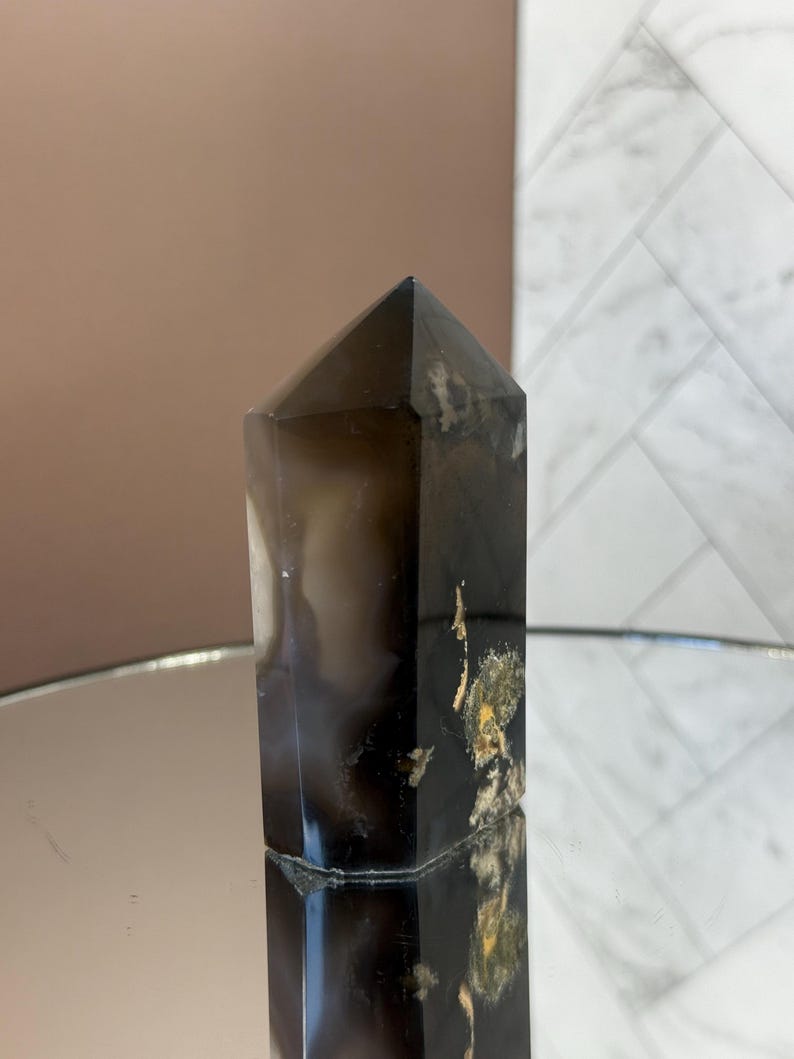 May include: A dark brown crystal obelisk with a pointed top. The crystal has a polished surface with areas of lighter color and some gold-colored inclusions. The obelisk is standing upright, reflecting on a mirrored surface.