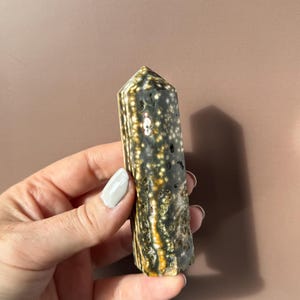 Puede incluir: Una gema pulida, de forma columnar, con un patr&oacute;n moteado de blanco, negro y dorado. El cristal se sostiene en una mano, mostrando sus tonos naturales y terrosos y su composici&oacute;n mineral &uacute;nica. La gema es un art&iacute;culo decorativo natural.