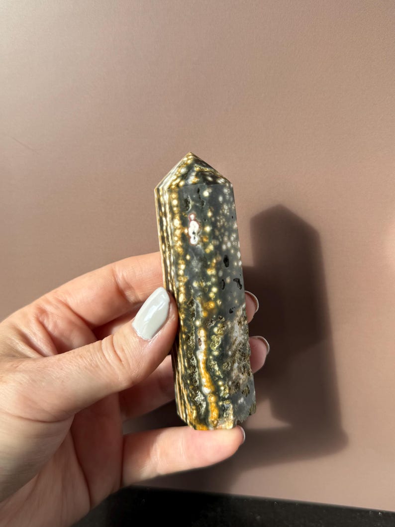 Puede incluir: Una gema pulida, de forma columnar, con un patr&oacute;n moteado de blanco, negro y dorado. El cristal se sostiene en una mano, mostrando sus tonos naturales y terrosos y su composici&oacute;n mineral &uacute;nica. La gema es un art&iacute;culo decorativo natural.