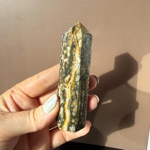 Puede incluir: Un obelisco de piedra preciosa pulida con una parte superior puntiaguda. La piedra presenta una mezcla de colores, incluyendo negro, blanco y dorado, con un patr&oacute;n moteado. El obelisco se sostiene en una mano sobre un fondo neutro.