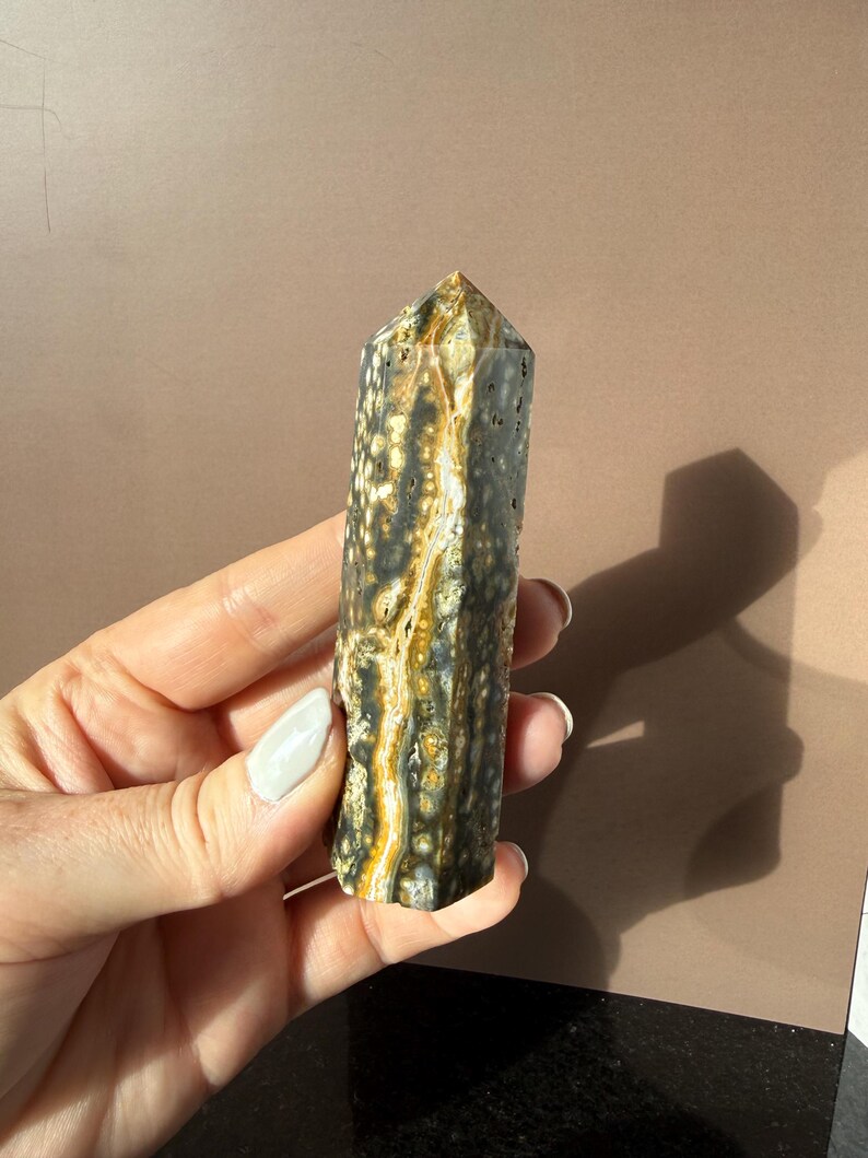 Puede incluir: Un obelisco de piedra preciosa pulida con una parte superior puntiaguda. La piedra presenta una mezcla de colores, incluyendo negro, blanco y dorado, con un patr&oacute;n moteado. El obelisco se sostiene en una mano sobre un fondo neutro.