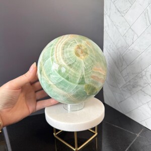 May include: A polished green onyx sphere featuring natural banded patterns in shades of light green, white, and soft orange. The stone orb rests on a clear circular stand, which is supported by a white marble base with a gold-toned metal frame.