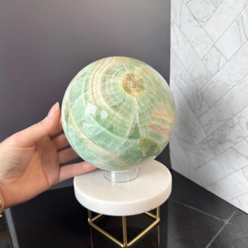 May include: A polished green onyx sphere featuring natural banded patterns in shades of light green, white, and soft orange. The stone orb rests on a clear circular stand, which is supported by a white marble base with a gold-toned metal frame.