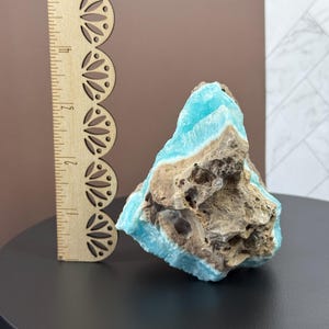 May include: A rough-cut, natural stone specimen with a striking contrast of colors. The stone features a vibrant turquoise hue on its surface, contrasted by a rough, brown matrix. A wooden ruler is placed next to the stone for scale.