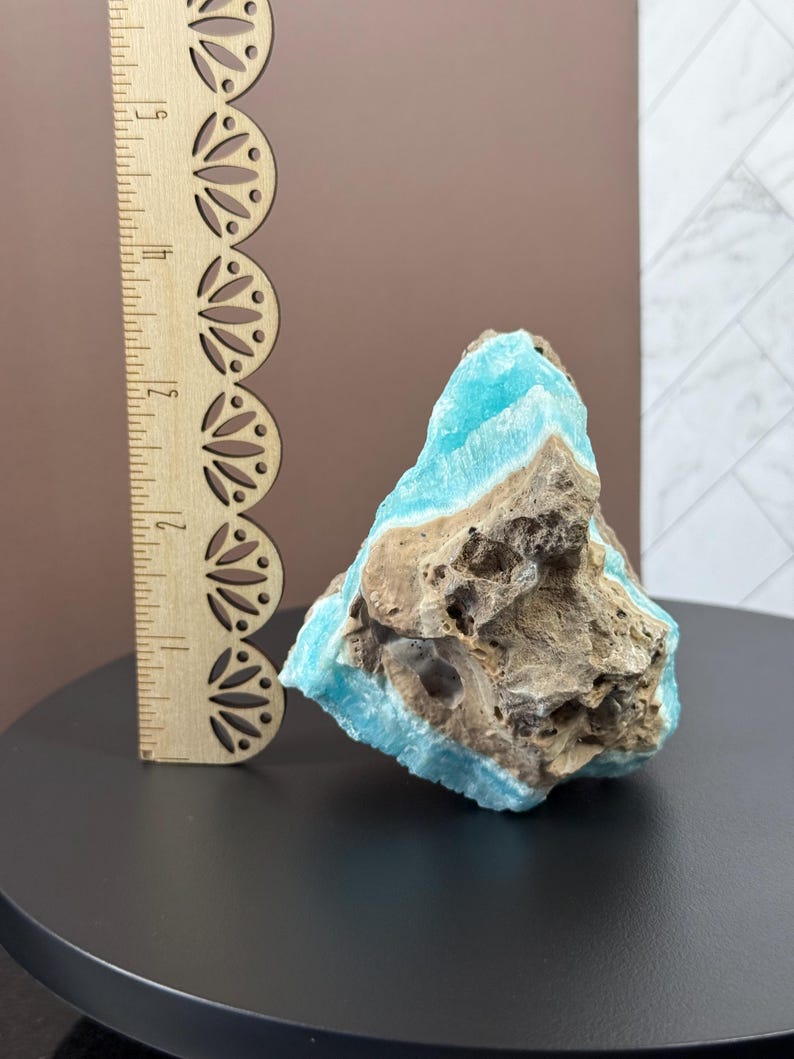 May include: A rough-cut, natural stone specimen with a striking contrast of colors. The stone features a vibrant turquoise hue on its surface, contrasted by a rough, brown matrix. A wooden ruler is placed next to the stone for scale.