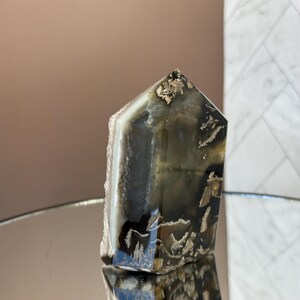 May include: A polished, rectangular crystal with a pointed top. The crystal features a mix of dark gray, white, and gold tones. The base has a rough, natural edge. The crystal is displayed on a reflective surface.