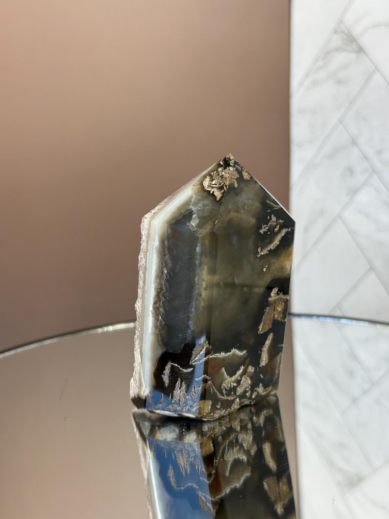 May include: A polished, rectangular crystal with a pointed top. The crystal features a mix of dark gray, white, and gold tones. The base has a rough, natural edge. The crystal is displayed on a reflective surface.