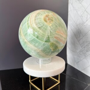 May include: A polished green onyx sphere featuring natural banded patterns in shades of light green, white, and soft orange. The sphere rests on a circular white stone base supported by a thin, square-shaped gold-toned metal frame stand.