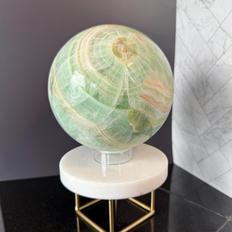 May include: A polished green onyx sphere featuring natural banded patterns in shades of light green, white, and soft orange. The sphere rests on a circular white stone base supported by a thin, square-shaped gold-toned metal frame stand.