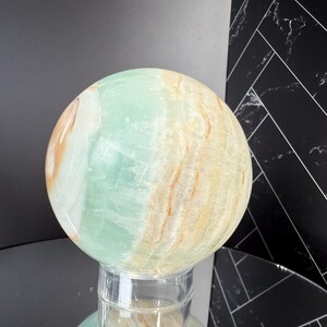 K&ouml;nnte beinhalten: Eine gro&szlig;e, polierte Kugel aus gr&uuml;nem und hellbraun geb&auml;ndertem Stein. Der Orb wird auf einem transparenten, zylindrischen St&auml;nder pr&auml;sentiert, der die Farben des Steins widerspiegelt. Die Oberfl&auml;che der Kugel hat ein marmoriertes Aussehen.