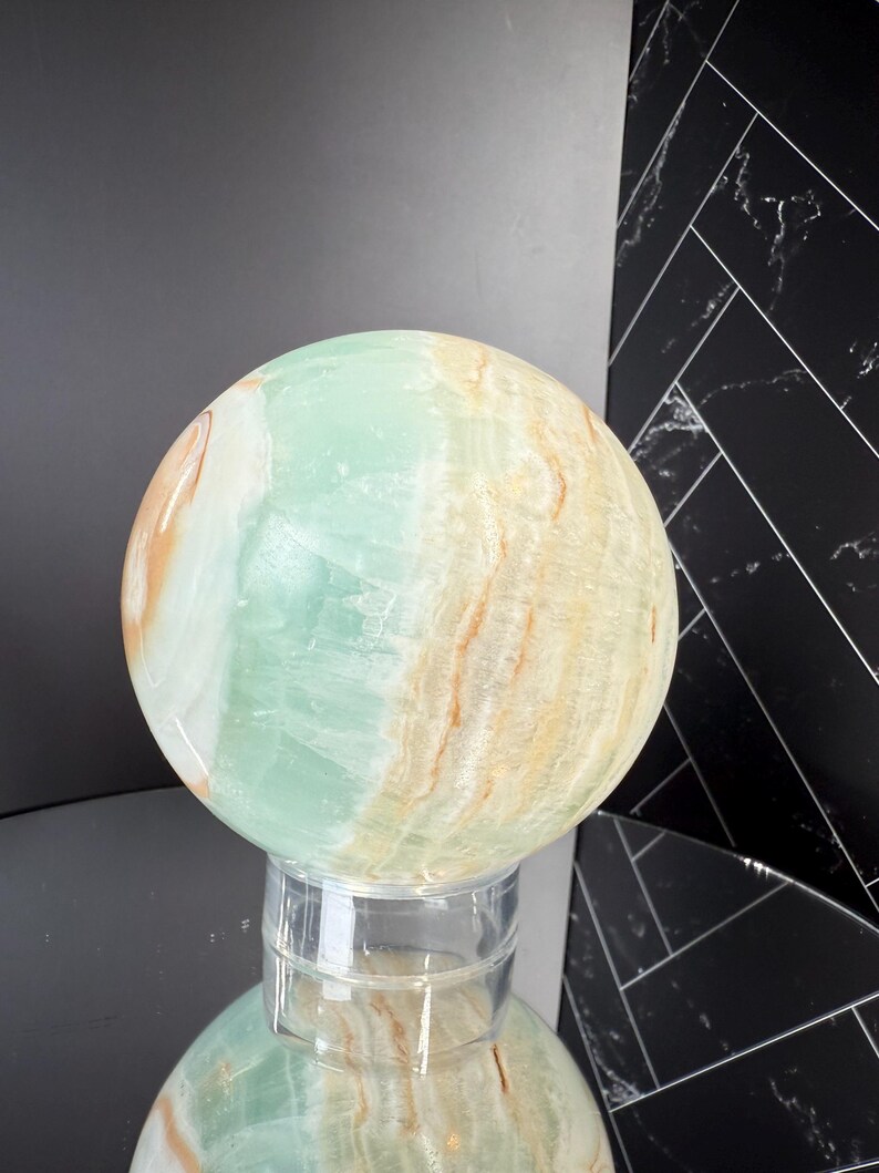 K&ouml;nnte beinhalten: Eine gro&szlig;e, polierte Kugel aus gr&uuml;nem und hellbraun geb&auml;ndertem Stein. Der Orb wird auf einem transparenten, zylindrischen St&auml;nder pr&auml;sentiert, der die Farben des Steins widerspiegelt. Die Oberfl&auml;che der Kugel hat ein marmoriertes Aussehen.