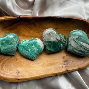 Puede incluir: Cuatro piedras pulidas en forma de corazón en tonos verdes y blancos están dispuestas en fila dentro de un cuenco de madera ovalado y poco profundo con un patrón de veta natural visible. Las piedras tienen superficies lisas y brillantes con texturas veteadas.