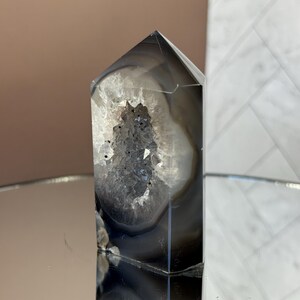 May include: A polished, gray and white agate crystal point with a hollowed-out center filled with sparkling, dark crystals. The crystal has a geometric, pointed top and is displayed on a reflective surface.