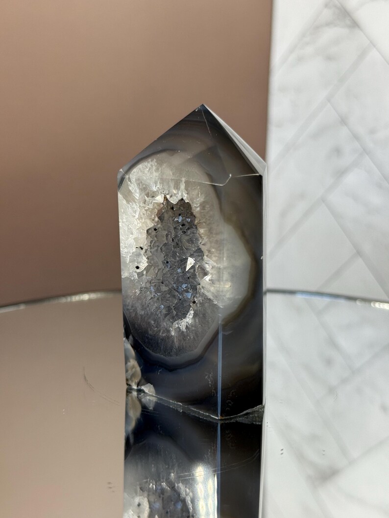 May include: A polished, gray and white agate crystal point with a hollowed-out center filled with sparkling, dark crystals. The crystal has a geometric, pointed top and is displayed on a reflective surface.