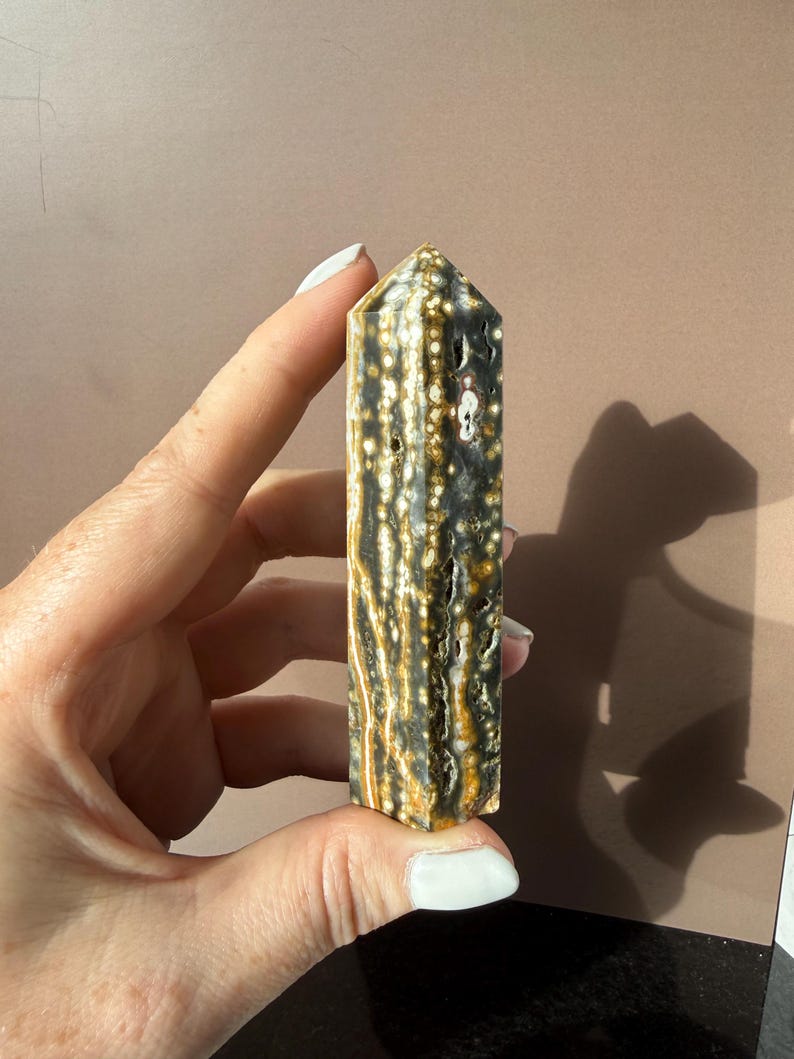 Puede incluir: Un obelisco de piedra preciosa rectangular y pulido. La piedra presenta un patr&oacute;n de manchas y vetas grises oscuras, crema y marr&oacute;n dorado. El obelisco se sostiene en una mano, mostrando su forma lisa y facetada y sus variaciones de color naturales.
