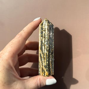 Puede incluir: Un cristal rectangular alto con una parte superior puntiaguda. El cristal es principalmente de color gris oscuro con un patr&oacute;n de puntos y vetas blancos y dorados. El cristal es sostenido por una mano con esmalte de u&ntilde;as blanco.