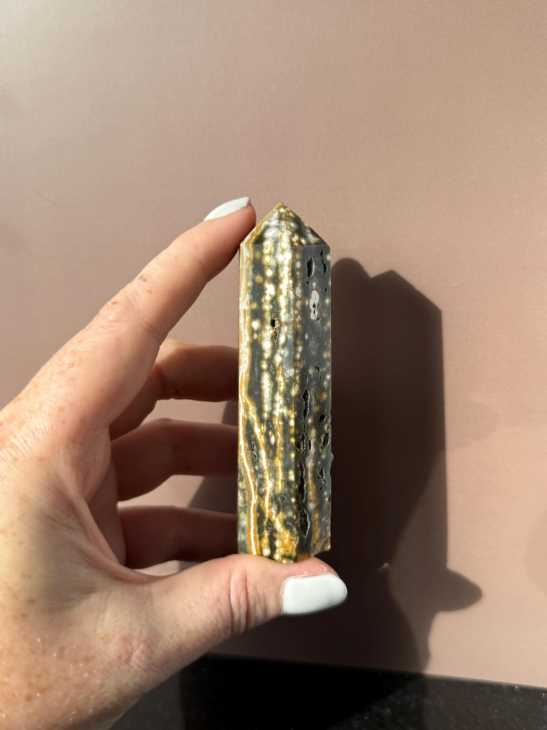 Puede incluir: Un cristal rectangular alto con una parte superior puntiaguda. El cristal es principalmente de color gris oscuro con un patr&oacute;n de puntos y vetas blancos y dorados. El cristal es sostenido por una mano con esmalte de u&ntilde;as blanco.