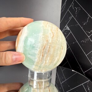 K&ouml;nnte beinhalten: Eine polierte Kugel aus gr&uuml;n-beigem Onyx, die auf einem transparenten Acrylst&auml;nder ruht. Die Oberfl&auml;che des Steins zeigt nat&uuml;rliche Muster und Farbvariationen, mit einer Mischung aus blassgr&uuml;nen und cremefarbenen T&ouml;nen. Die Kugel wird von einer Hand gehalten.