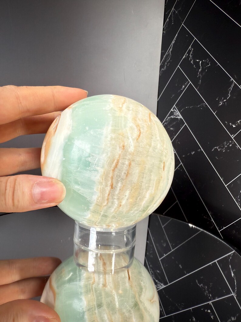 K&ouml;nnte beinhalten: Eine polierte Kugel aus gr&uuml;n-beigem Onyx, die auf einem transparenten Acrylst&auml;nder ruht. Die Oberfl&auml;che des Steins zeigt nat&uuml;rliche Muster und Farbvariationen, mit einer Mischung aus blassgr&uuml;nen und cremefarbenen T&ouml;nen. Die Kugel wird von einer Hand gehalten.