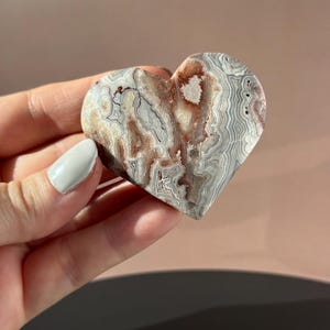 May include: A heart-shaped stone carving with a unique pattern of gray, white, and reddish-brown swirls. The polished surface of the stone is smooth, and the heart is held in a hand. The stone appears to be a decorative item.