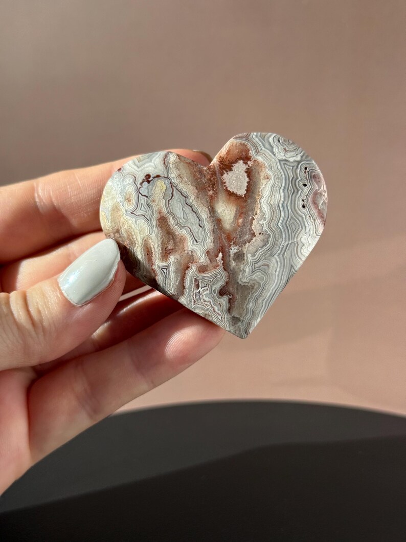 May include: A heart-shaped stone carving with a unique pattern of gray, white, and reddish-brown swirls. The polished surface of the stone is smooth, and the heart is held in a hand. The stone appears to be a decorative item.