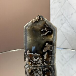 May include: A polished, hexagonal crystal tower with a pointed top. The stone features a mix of dark brown, black, and cream colors with a reflective surface. The crystal is displayed on a mirrored surface.